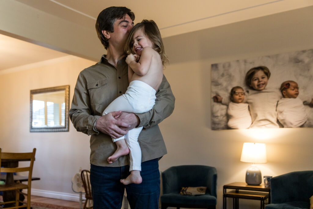 Dad kissing smiling daughter, dressed in tights, during in home documentary photo session.