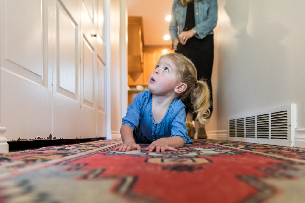 Toddler girl crawling on rug, looking up, in hallway on red rug with mom chasing her.