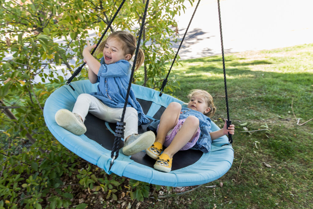 Twin girls laughing and swinging on round swing in their front yard during a Denver photo session.