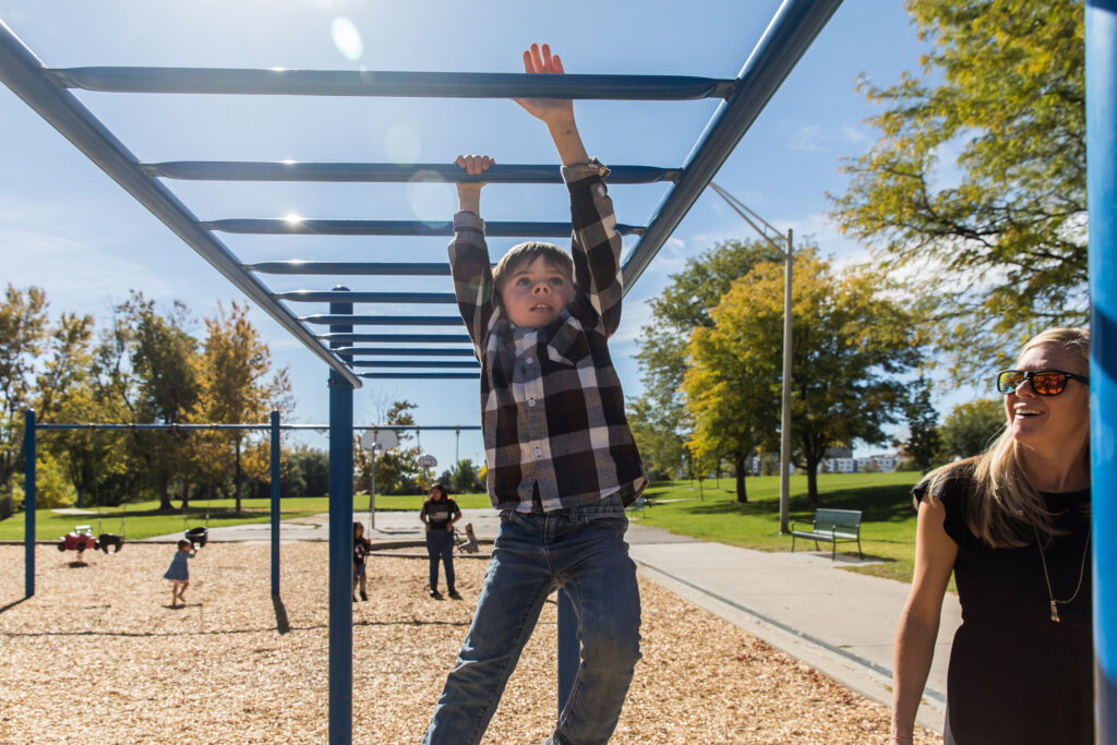 Boy going across the monkey bars in a park while his mother smiles and watches in Denver, Colorado.