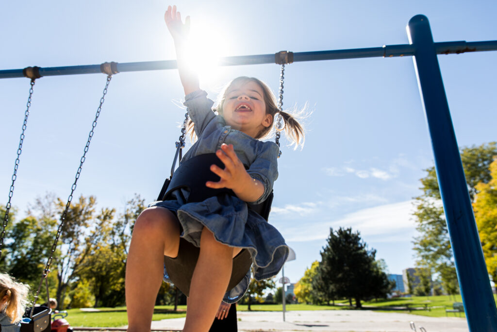 Toddler girl in pigtails smiling on a swing and reaching up to the sky.