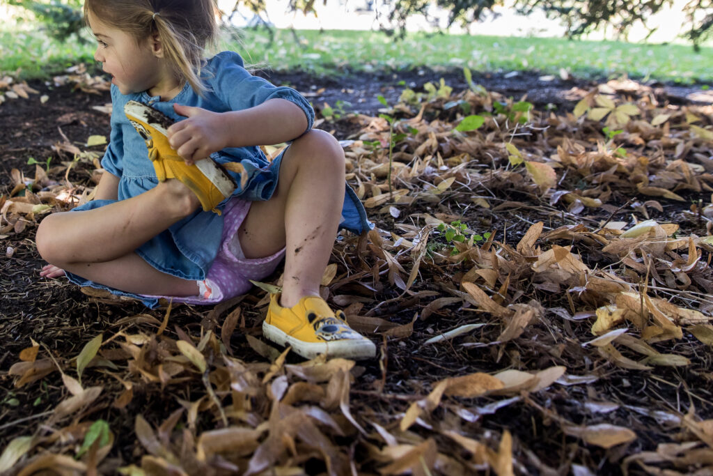 Toddler girl sitting in muddy leaves with dirty yellow shoes at a park in Denver, Colorado.