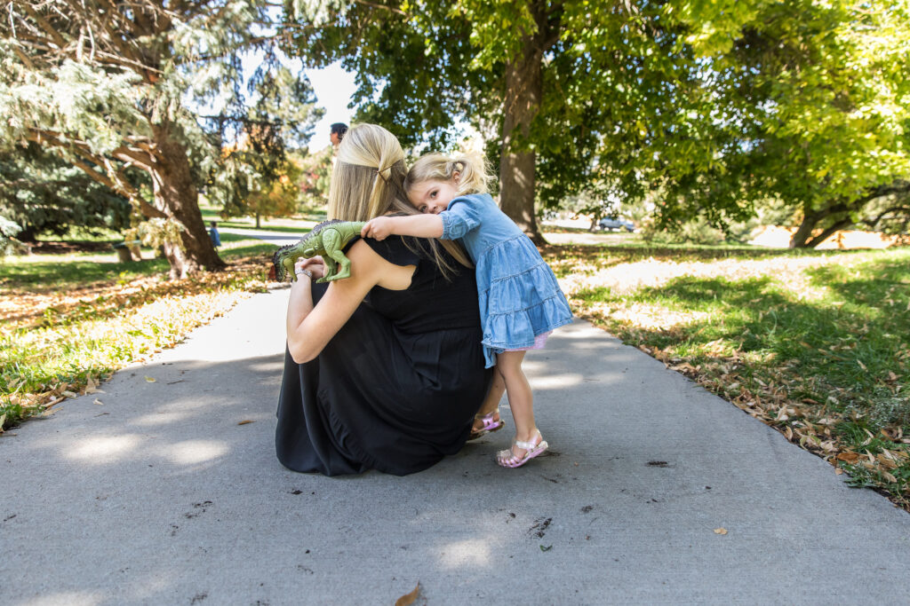 Toddler girl on tiptoes, hugging mom's back while holding large green toy dinosaur.