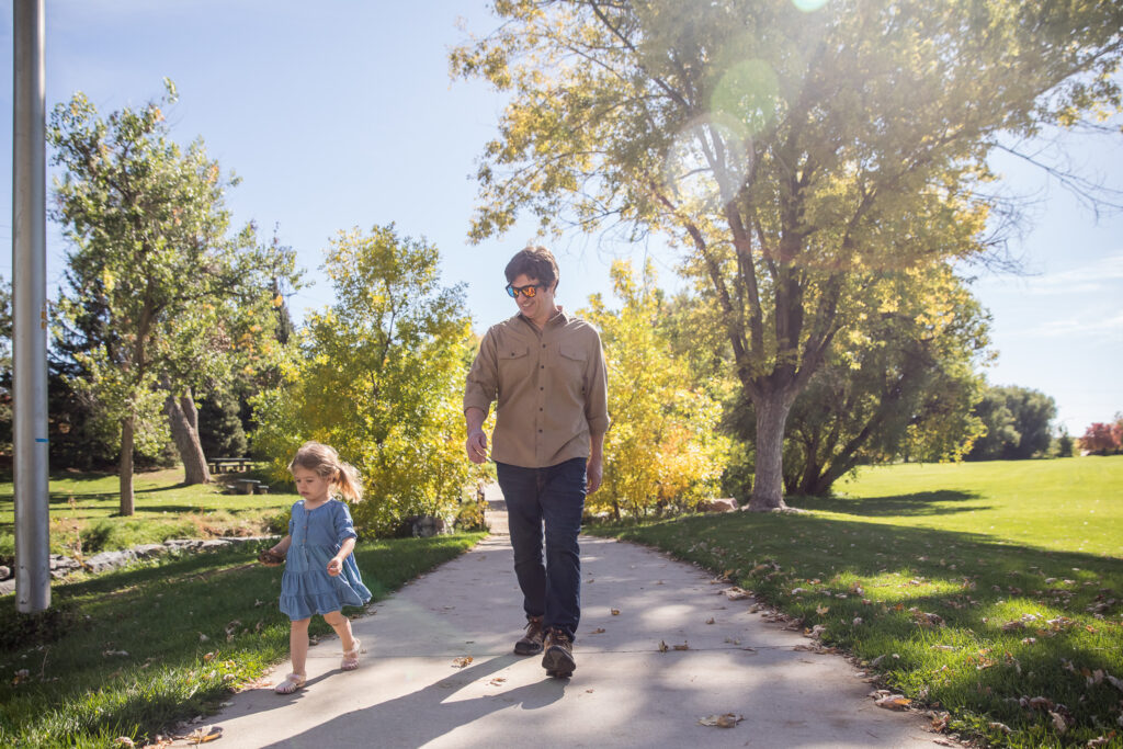 Dad chasing daughter down a tree-lined path through a park in Denver. CO.